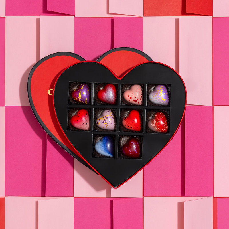 10 gorgeous heart-shaped chocolates in a variety of pink, purple and blue colors. The chocolates are displayed in a red heart-shaped gift box on top of a light and dark pink background. 